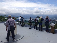 展望台から秩父盆地を見下ろしている10名弱の参加者の後ろ姿。雲が多いが晴れ始めている。